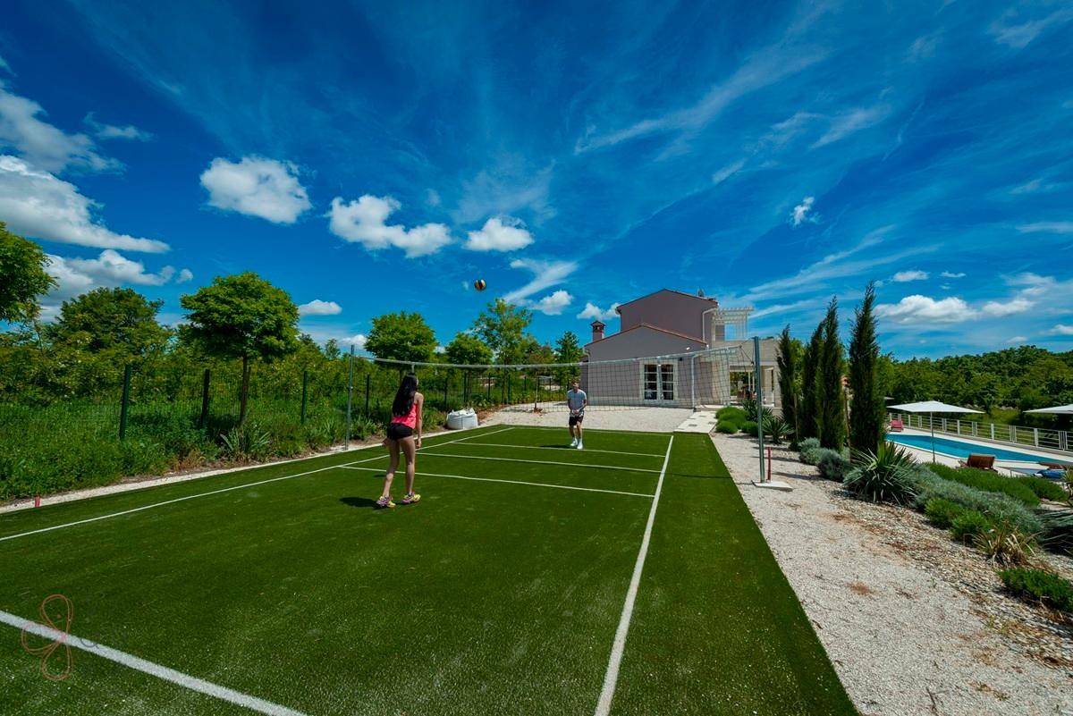 Volleyballfeld im Freien mit Spielern, im Hintergrund eine Villa mit Poolbereich.