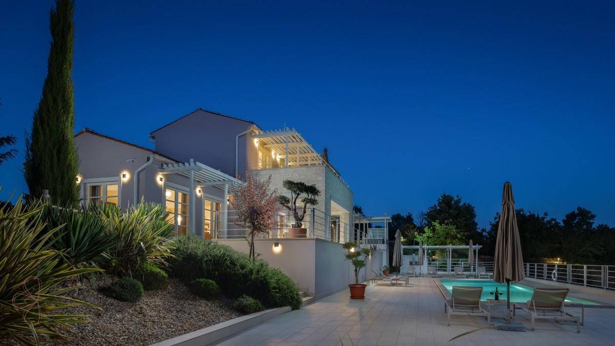Seitliche Ansicht einer luxuriösen Villa mit Pool, gepflegtem Garten und stimmungsvoller Beleuchtung.