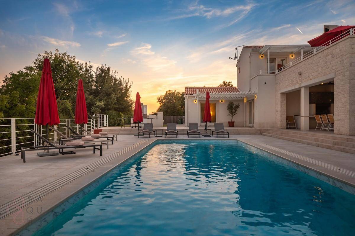 Abendliche Ansicht des Hauses mit beleuchtetem Poolbereich und roten Sonnenschirmen bei Sonnenuntergang.