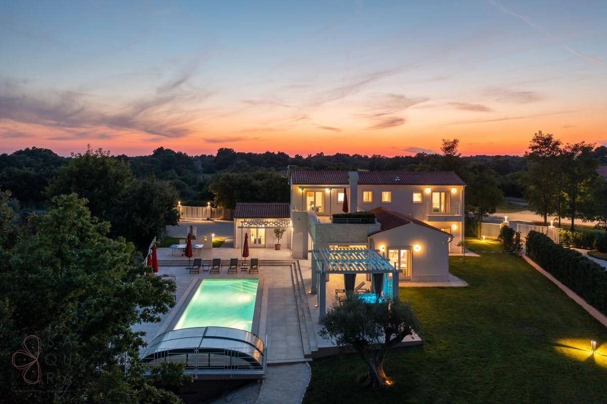 Beeindruckende Villa mit beleuchtetem Poolbereich und weitläufigem Garten bei Sonnenuntergang.