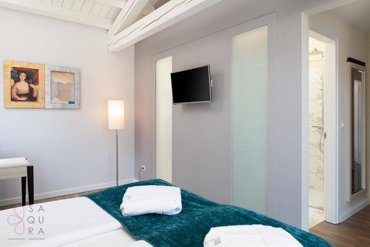 Schlafzimmer mit Holzbalkendecke, Wandfernseher und direktem Zugang zum Badezimmer.