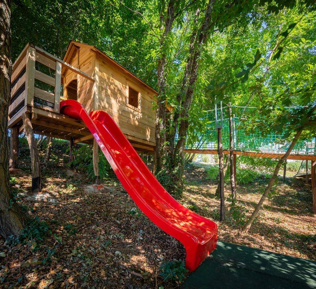 Baumhaus mit roter Rutsche in einem grünen, bewaldeten Spielbereich für Kinder.
