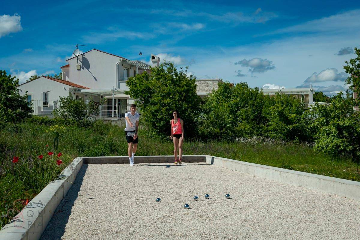 Außenbereich mit Boccia-Platz und Grünflächen, umgeben von Bäumen und einem Haus.