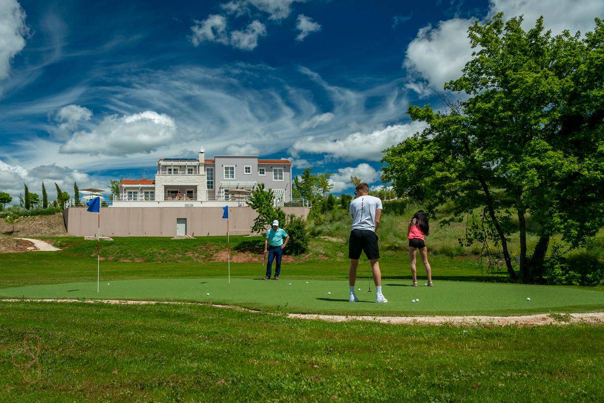 Personen beim Golfspielen auf dem Putting-Green mit einer luxuriösen Villa im Hintergrund unter blauem Himmel.
