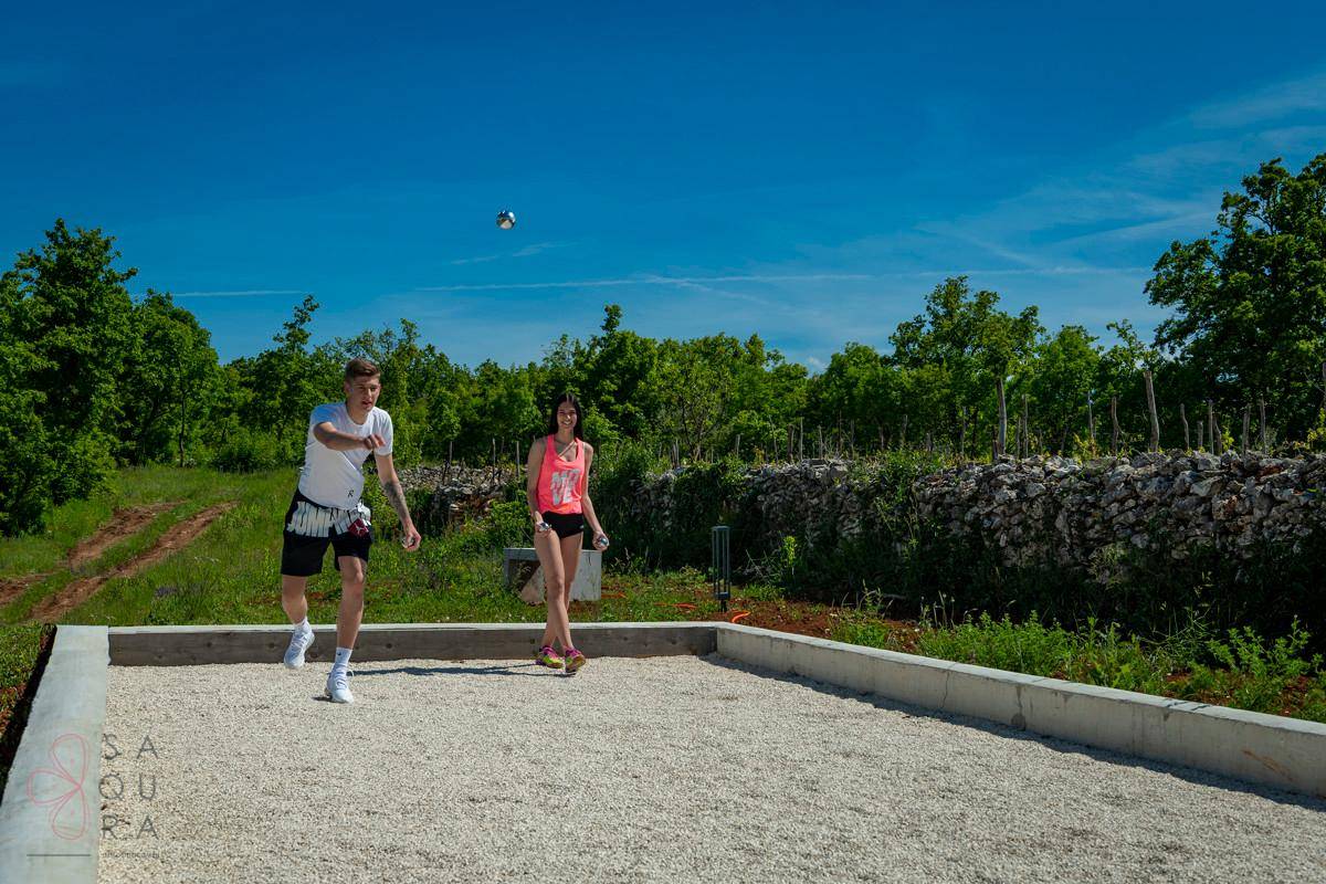 Zwei Personen spielen Boccia auf einem Kiesplatz, umgeben von grüner Landschaft unter blauem Himmel.