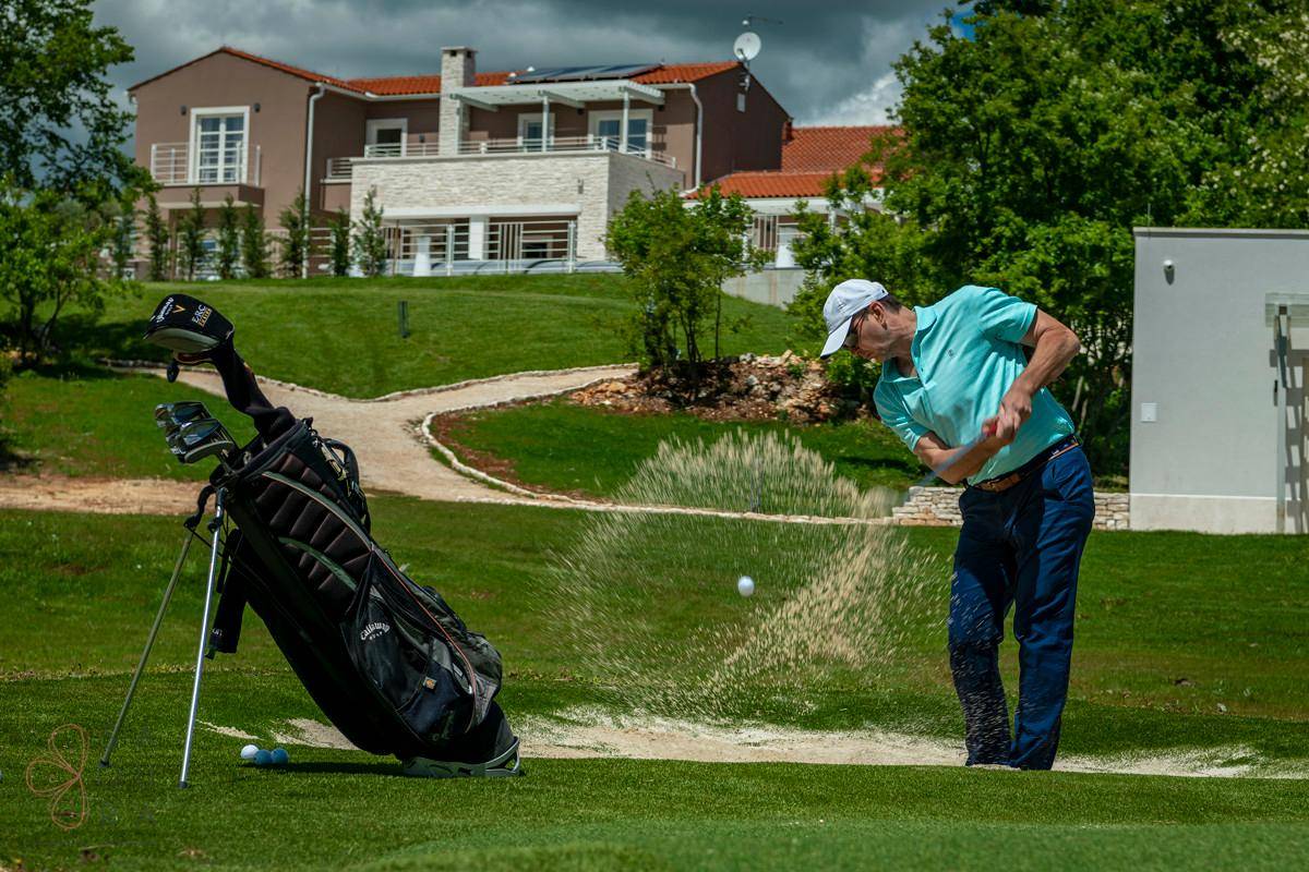 Ein Golfer schlägt einen Ball aus einem Sandbunker, mit einer Villa und grünen Hügeln im Hintergrund.