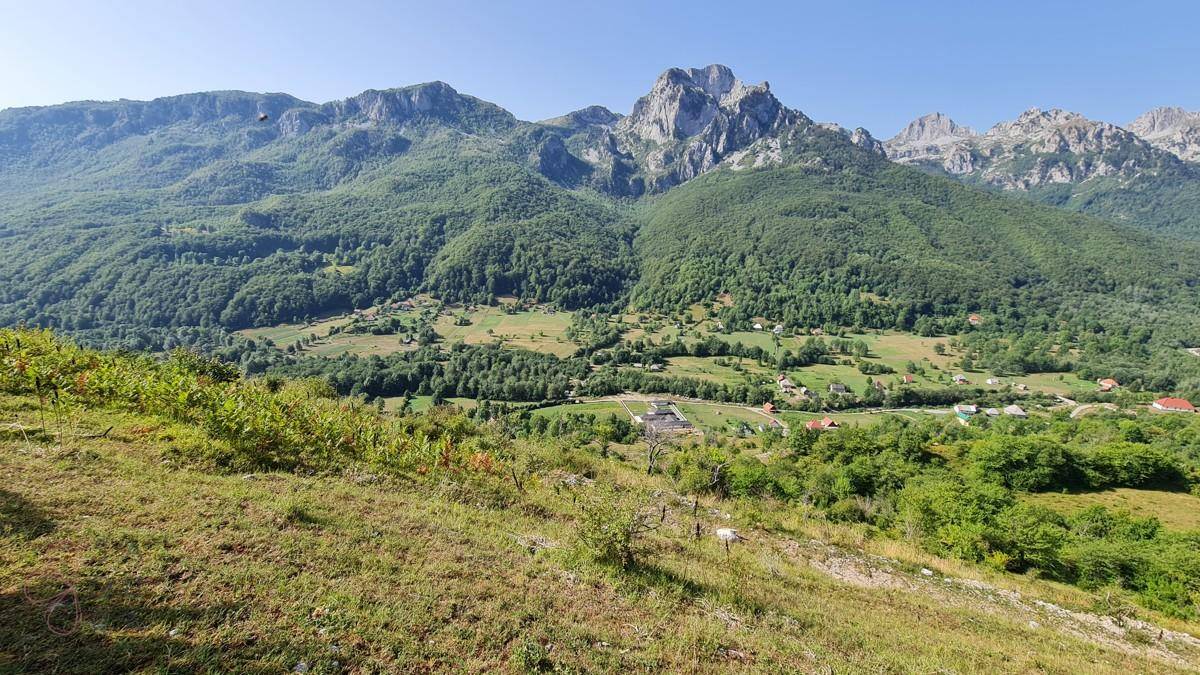 Panoramablick auf eine weite, grüne Berglandschaft mit Tälern und kleinen Siedlungen unter blauem Himmel.