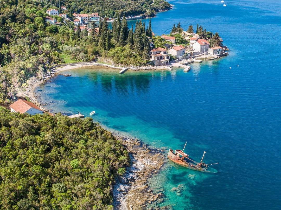 Luftaufnahme einer idyllischen Bucht mit klarem Wasser, grünen Hügeln und einem Schiffswrack am Ufer.