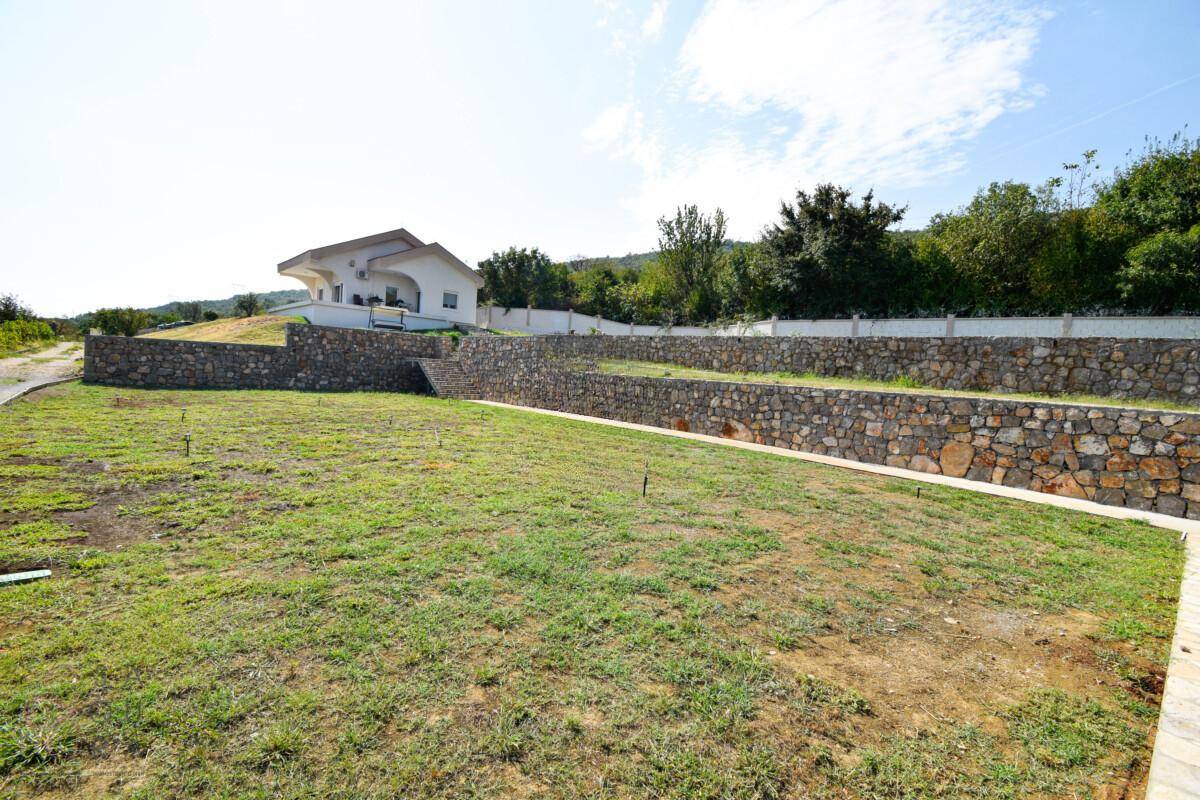 Weitläufige Rasenfläche mit Natursteinmauern und dem Haus im Hintergrund, unter blauem Himmel.