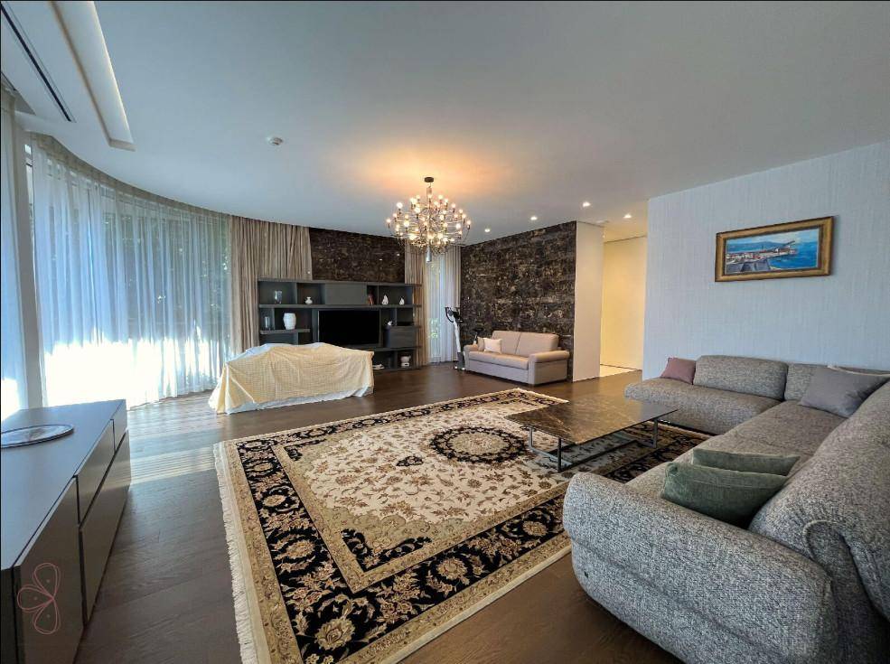 Geräumiges Wohnzimmer mit großem Sofa, elegantem Teppich, dunklem Holzboden und raumhohen Fenstern mit Vorhängen.