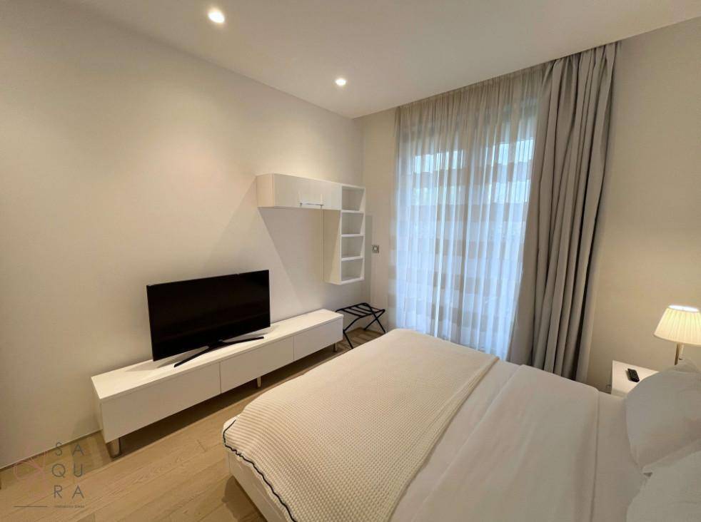 Schlafzimmer mit weißem Bett, Fernseher auf einem Sideboard und großem Fenster mit Vorhängen.