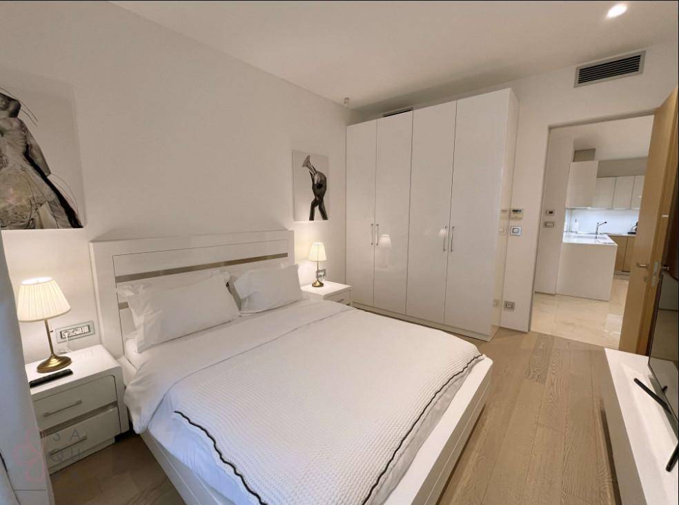 Schlafzimmer mit weißem Bett, Nachttischen und großem Einbauschrank, mit Blick zur Küche.
