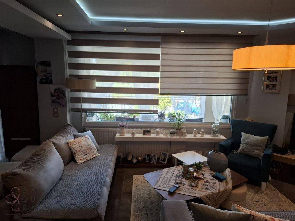 Gemütliches Wohnzimmer mit beigem Sofa, blauem Sessel und Fenster mit modernen Jalousien.