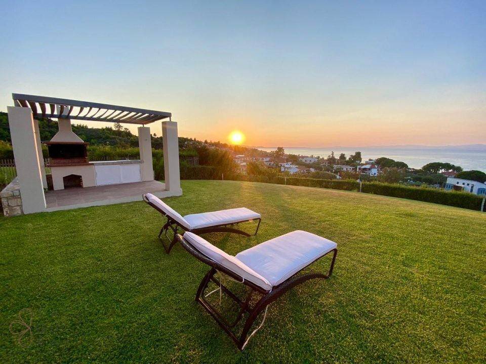 Gepflegter Rasenbereich mit Sonnenliegen und Grillplatz bei Sonnenuntergang mit Meerblick.