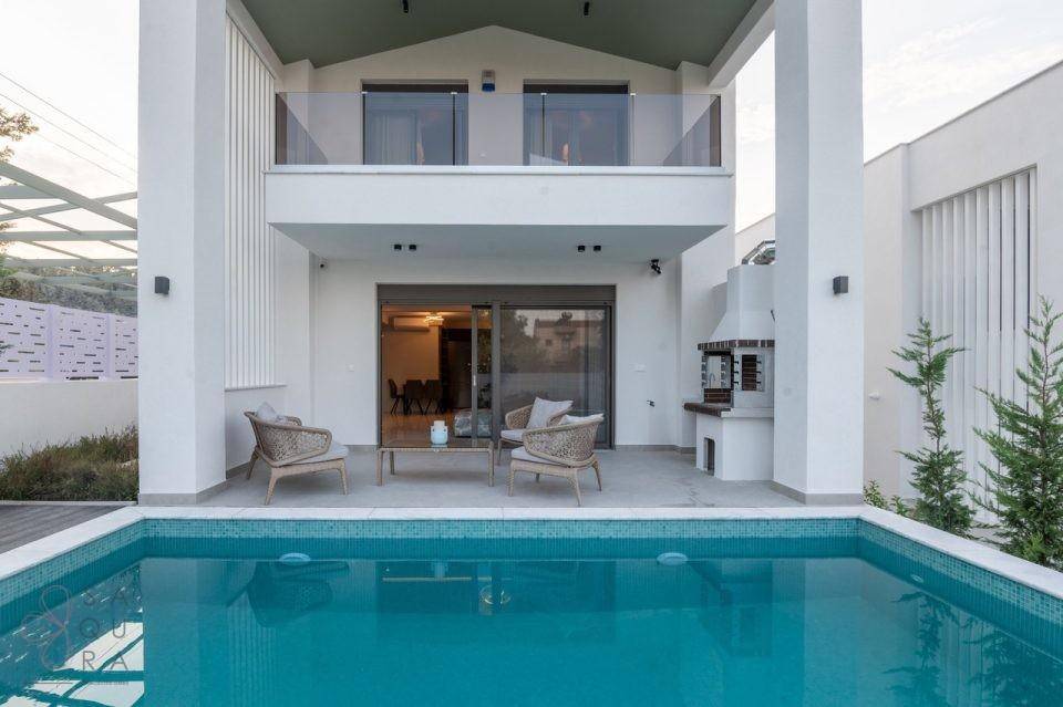 Ansicht der modernen Villa von vorne mit großem Pool und Balkon im Obergeschoss.