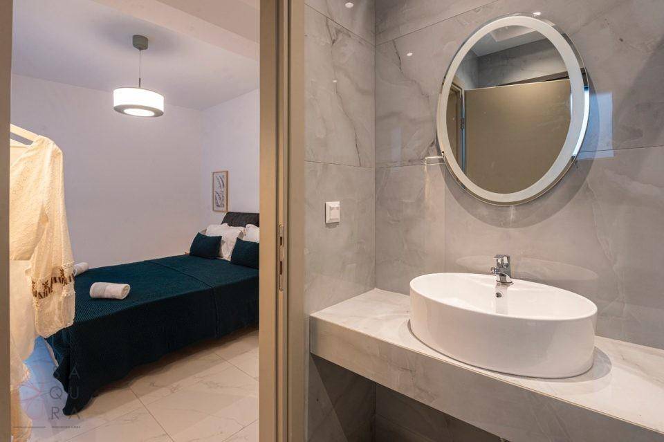 Modernes Badezimmer mit ovalem Waschbecken, beleuchtetem Spiegel und Blick ins Schlafzimmer.