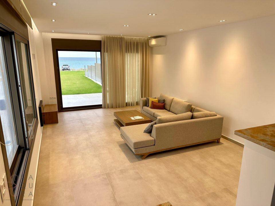 Helles Wohnzimmer mit beigem Sofa, Fliesenboden und direktem Zugang zum Garten mit Meerblick.