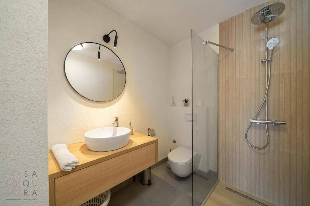 Helles Badezimmer mit modernem Waschtisch, rundem Spiegel und ebenerdiger Glasdusche.