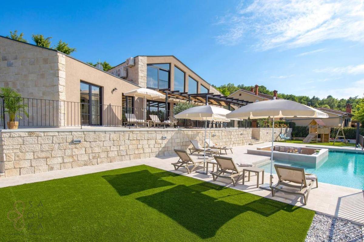 Moderne Villa mit großem Pool, Liegebereich und Kunstrasenfläche unter blauem Himmel.
