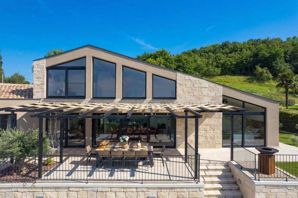 Moderne Villa mit großer Terrasse unter einer Pergola und weiten Fenstern zum Wohnbereich.