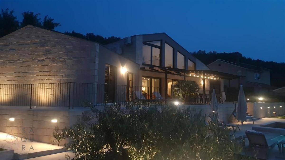 Moderne Villa bei Nacht, beleuchtet mit Terrasse, Pergola und Poolbereich, umgeben von Natur.