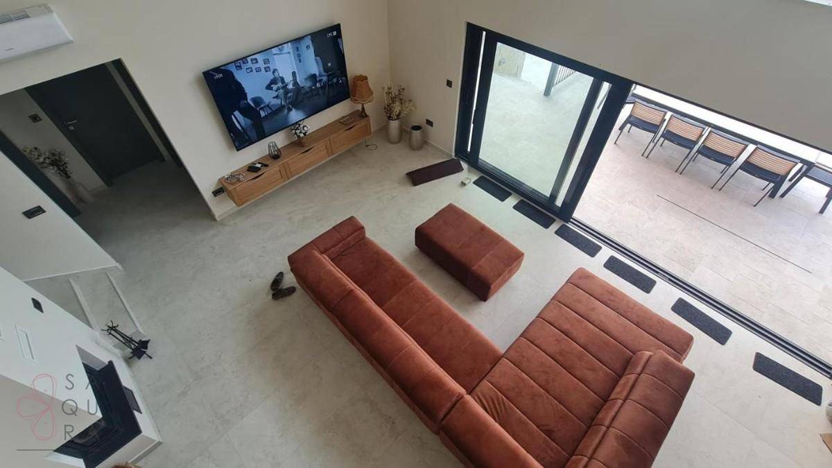 Heller Wohnbereich von oben gesehen mit großem Sofa, Wandfernseher und Zugang zu einer Außenterrasse.