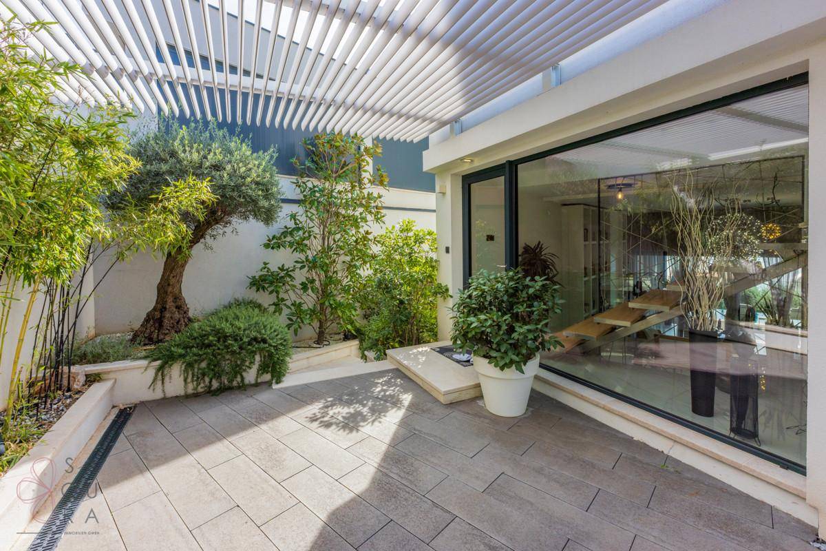 Helle Terrasse mit mediterraner Bepflanzung und großzügigen Glasfronten zum Innenbereich.