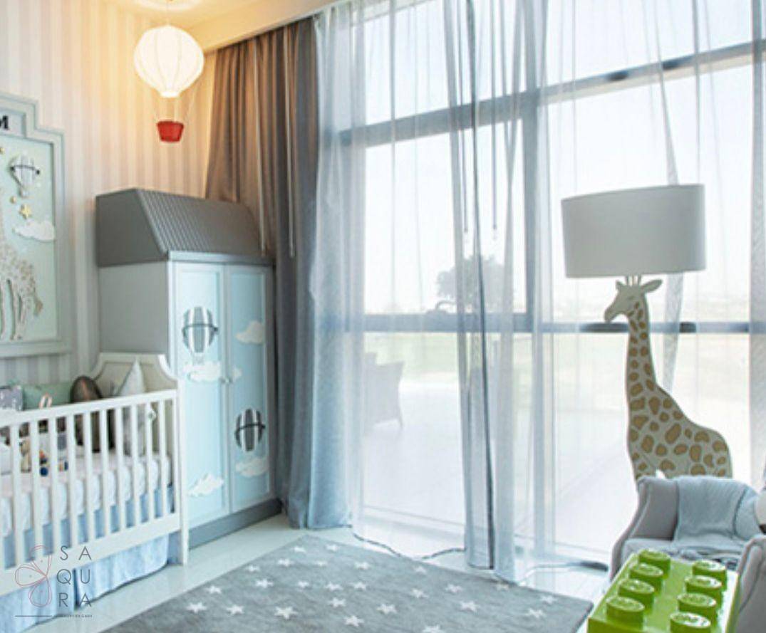 Helles Kinderzimmer mit weißem Babybett, blauem Kleiderschrank und großen Fenstern mit transparenten Vorhängen.
