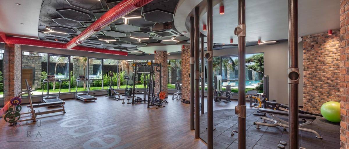 Modernes Fitnessstudio mit vielfältigen Geräten, Holzboden und großen Fenstern mit Blick auf den Poolbereich.