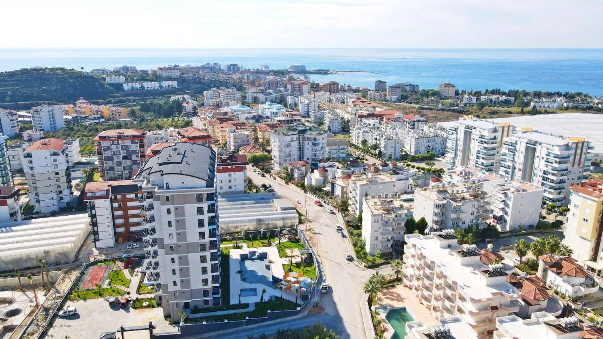 Weitläufige Stadtansicht mit zahlreichen Gebäuden, Grünflächen und dem Meer im Hintergrund.