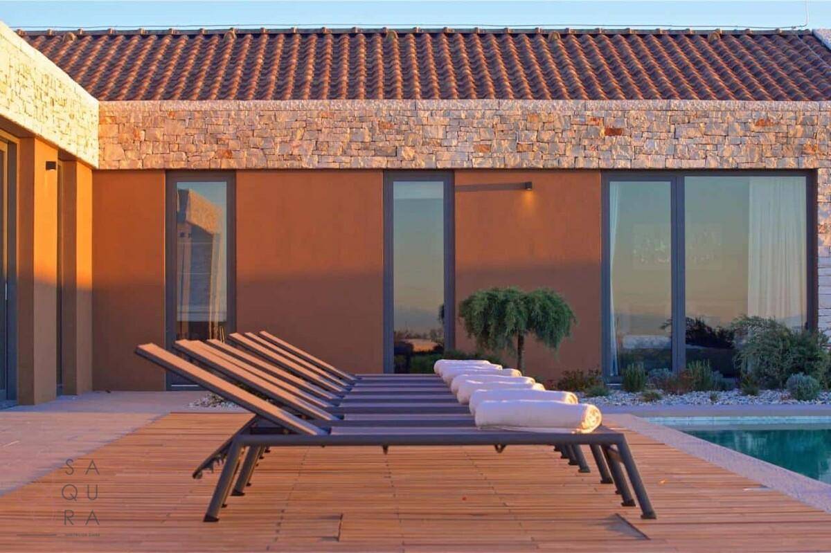 Sonnenliegen auf dem Holzdeck am Pool, mit der Villa im warmen Licht des Sonnenuntergangs.