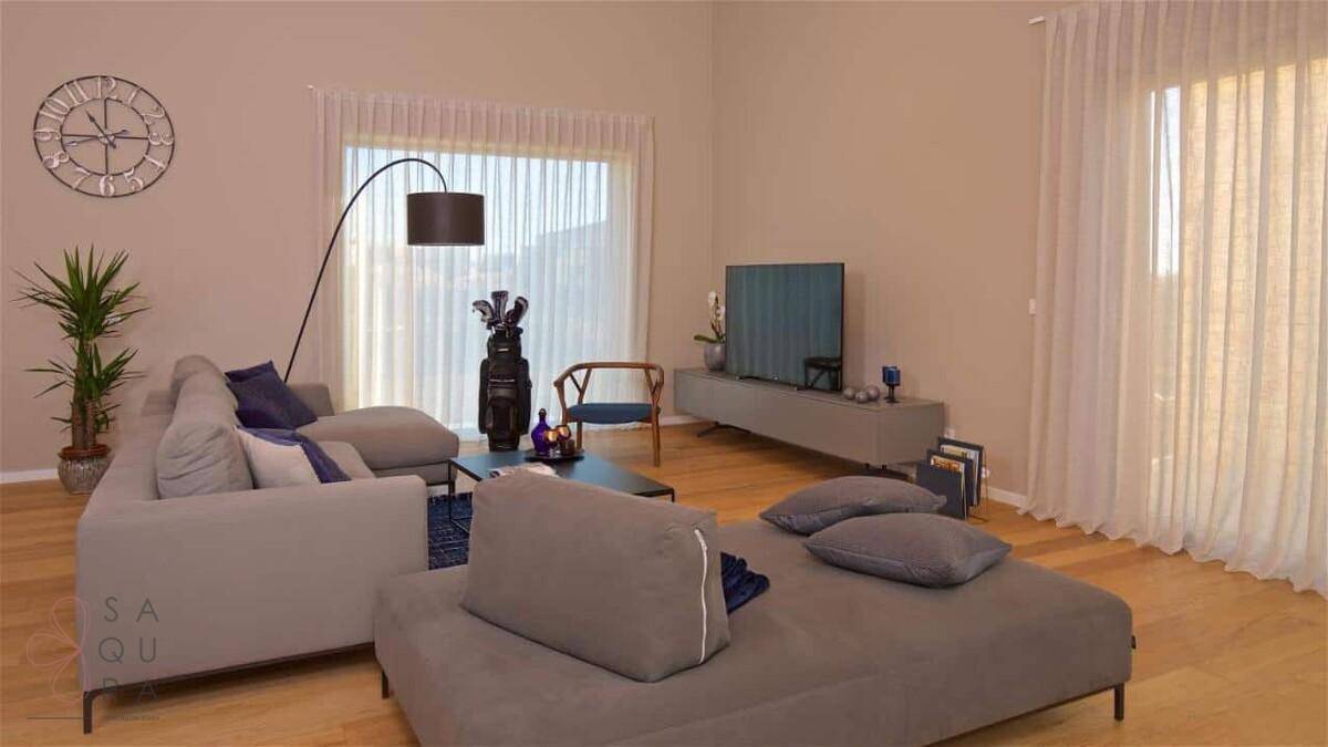 Komfortables Wohnzimmer mit großem Sofa, Flachbildfernseher und hellen Fenstern mit Vorhängen.