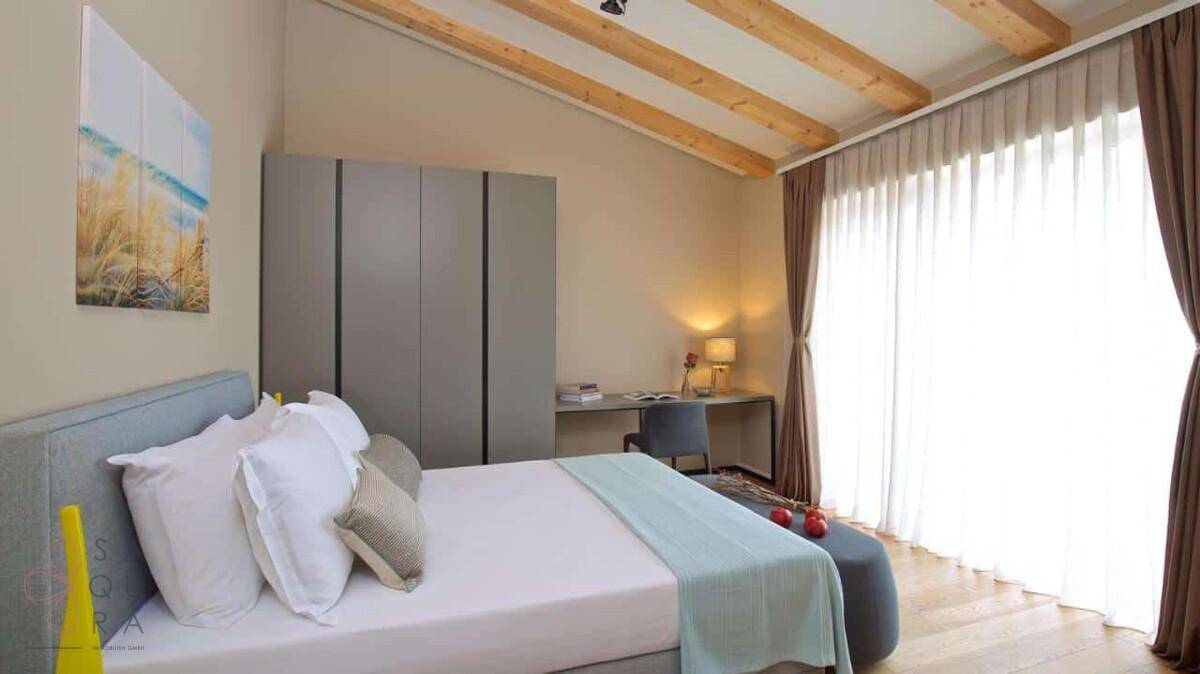 Helles Schlafzimmer mit Holzbalkendecke, Schreibtisch und großem Fenster mit Vorhängen.