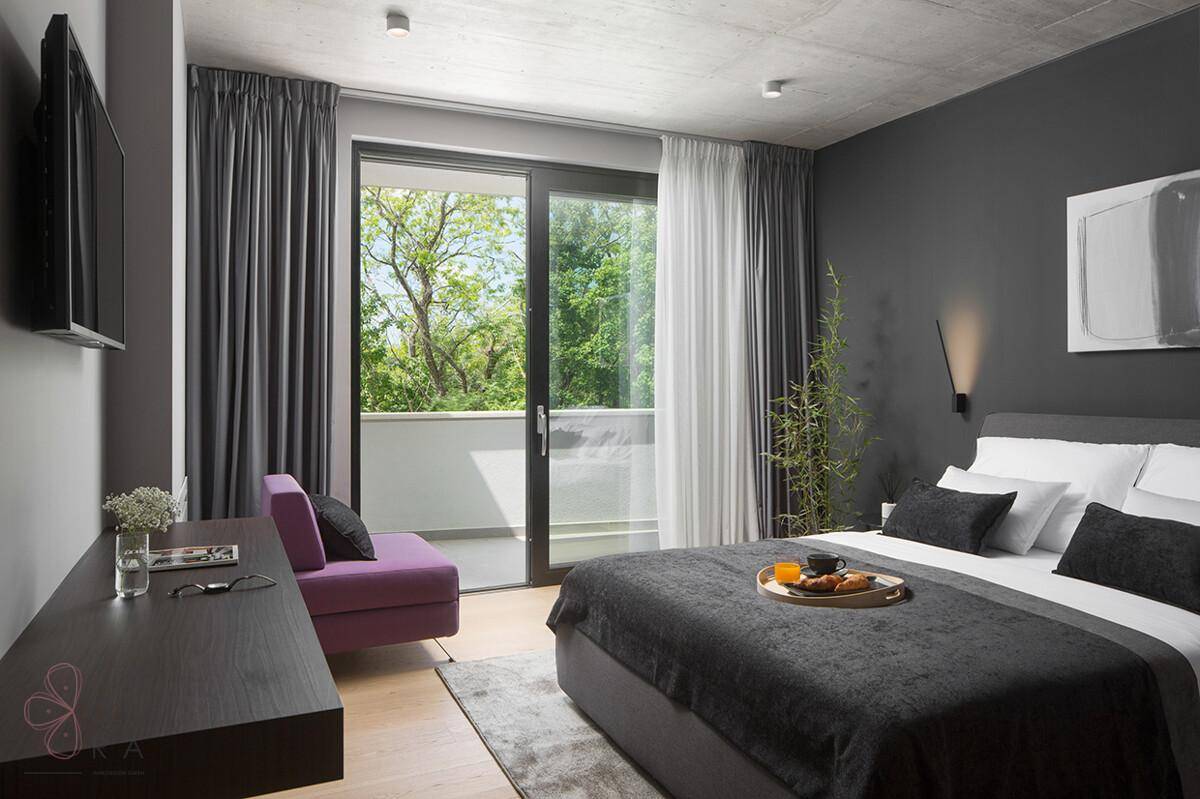 Helles Schlafzimmer mit Doppelbett, Schreibtisch und Balkonzugang mit Blick ins Grüne.
