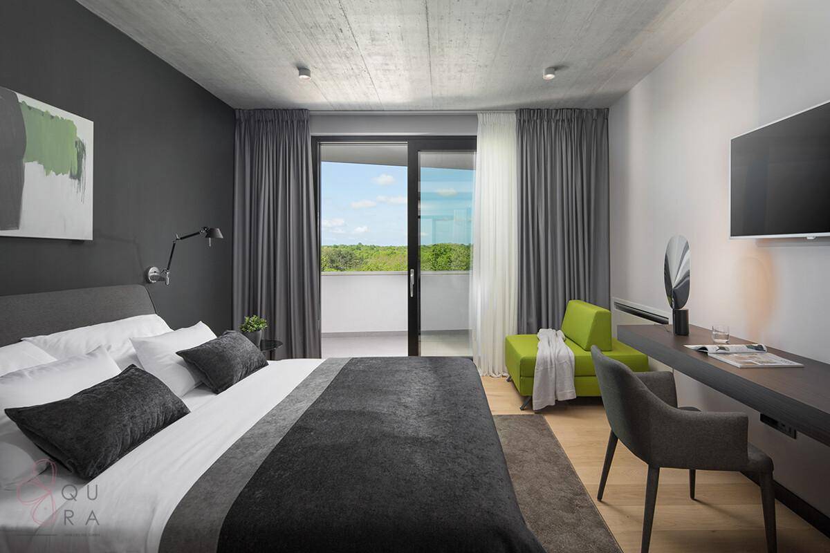 Modernes Schlafzimmer mit Doppelbett, Schreibtisch und Zugang zu einem Balkon mit grüner Aussicht.