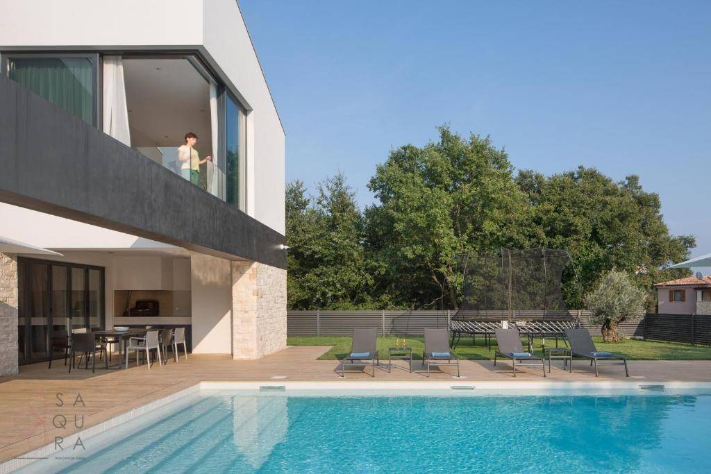 Weitläufiger Gartenbereich mit Pool, Liegestühlen und einer Person auf dem Balkon der modernen Villa.