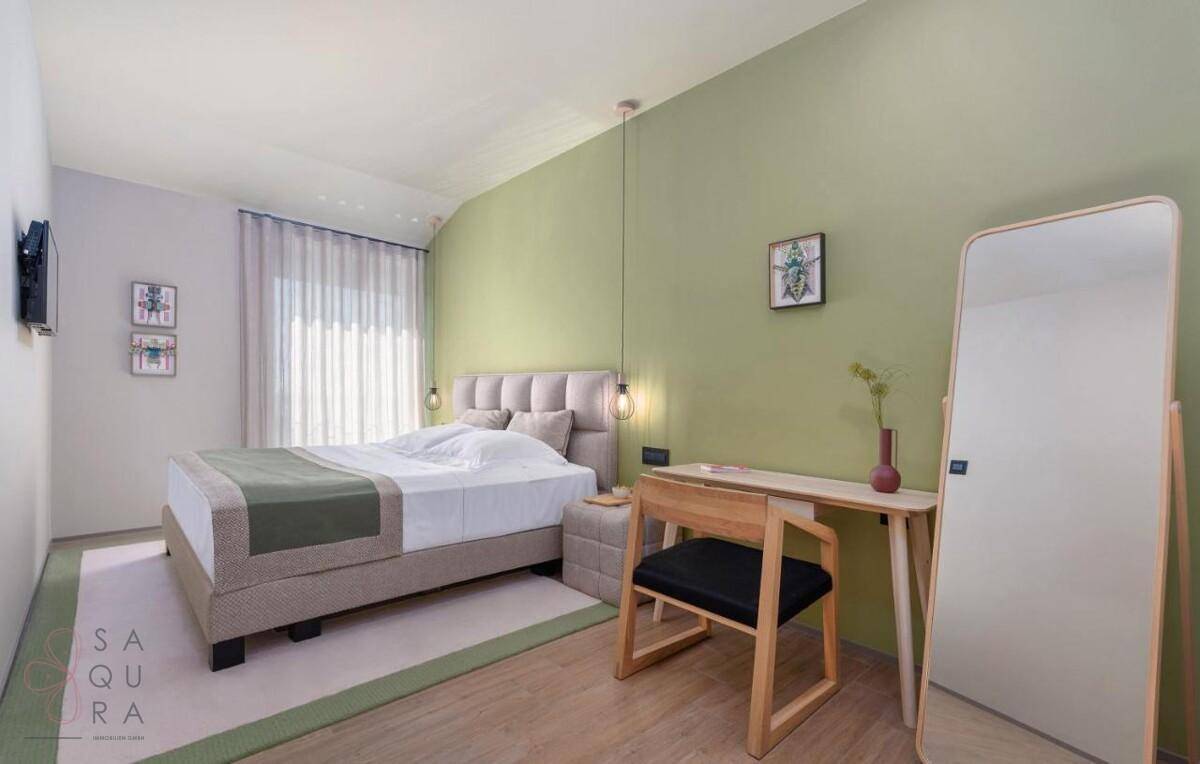 Helles Schlafzimmer mit Doppelbett, Schreibtisch und grünen Akzenten an der Wand.
