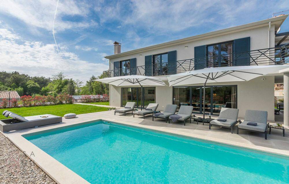 Moderne Hausfassade mit großem Pool, Liegebereich und gepflegtem Garten unter blauem Himmel.