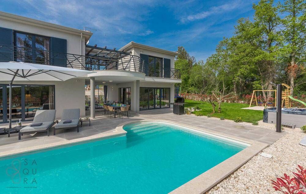 Gesamtansicht der Villa mit geschwungenem Pool, Whirlpool, Kinderspielplatz und üppigem Grün.