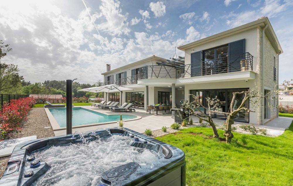 Moderne Villa mit großem Pool und sprudelndem Whirlpool im Vordergrund, umgeben von Grün.