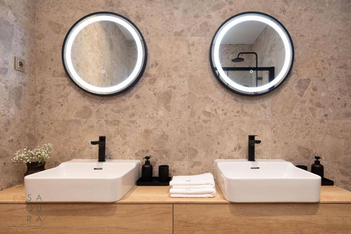 Modernes Badezimmer mit Doppelwaschbecken, runden beleuchteten Spiegeln und eleganten Fliesen.