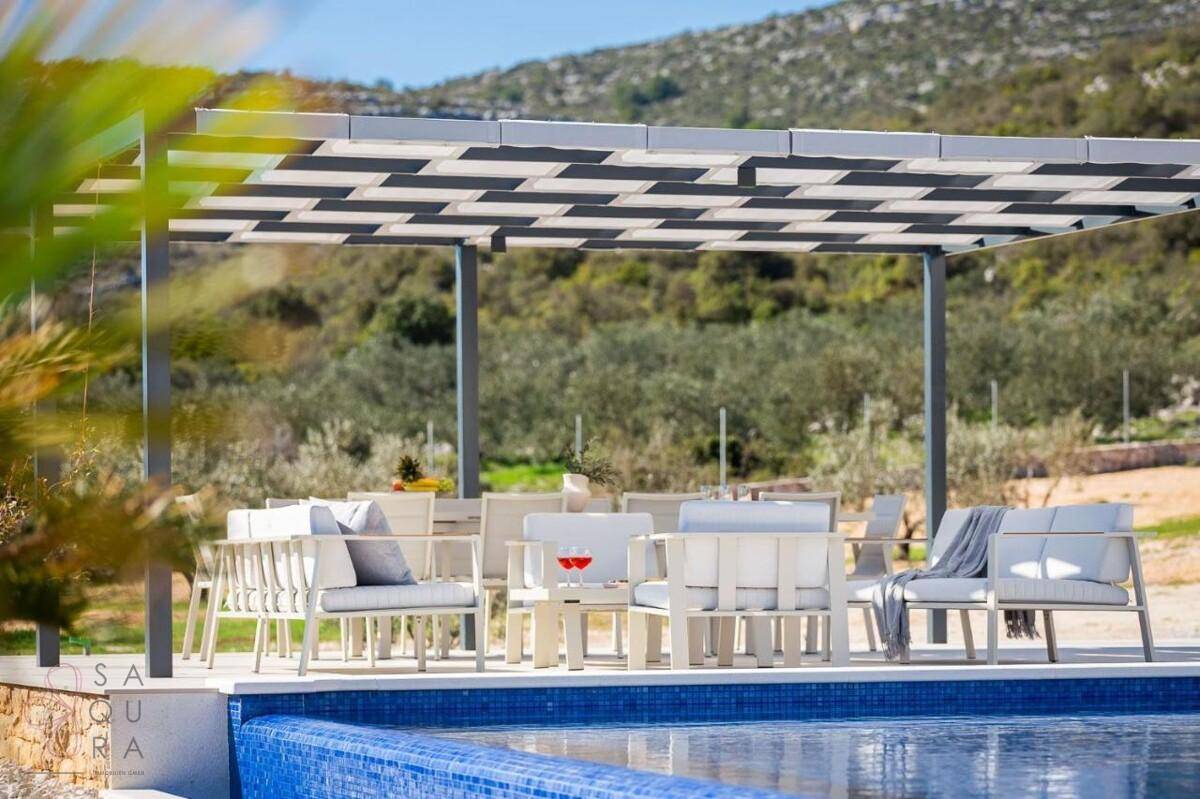 Stilvoller Essbereich im Freien unter einer Pergola, direkt am Pool mit Blick auf die grüne Umgebung.