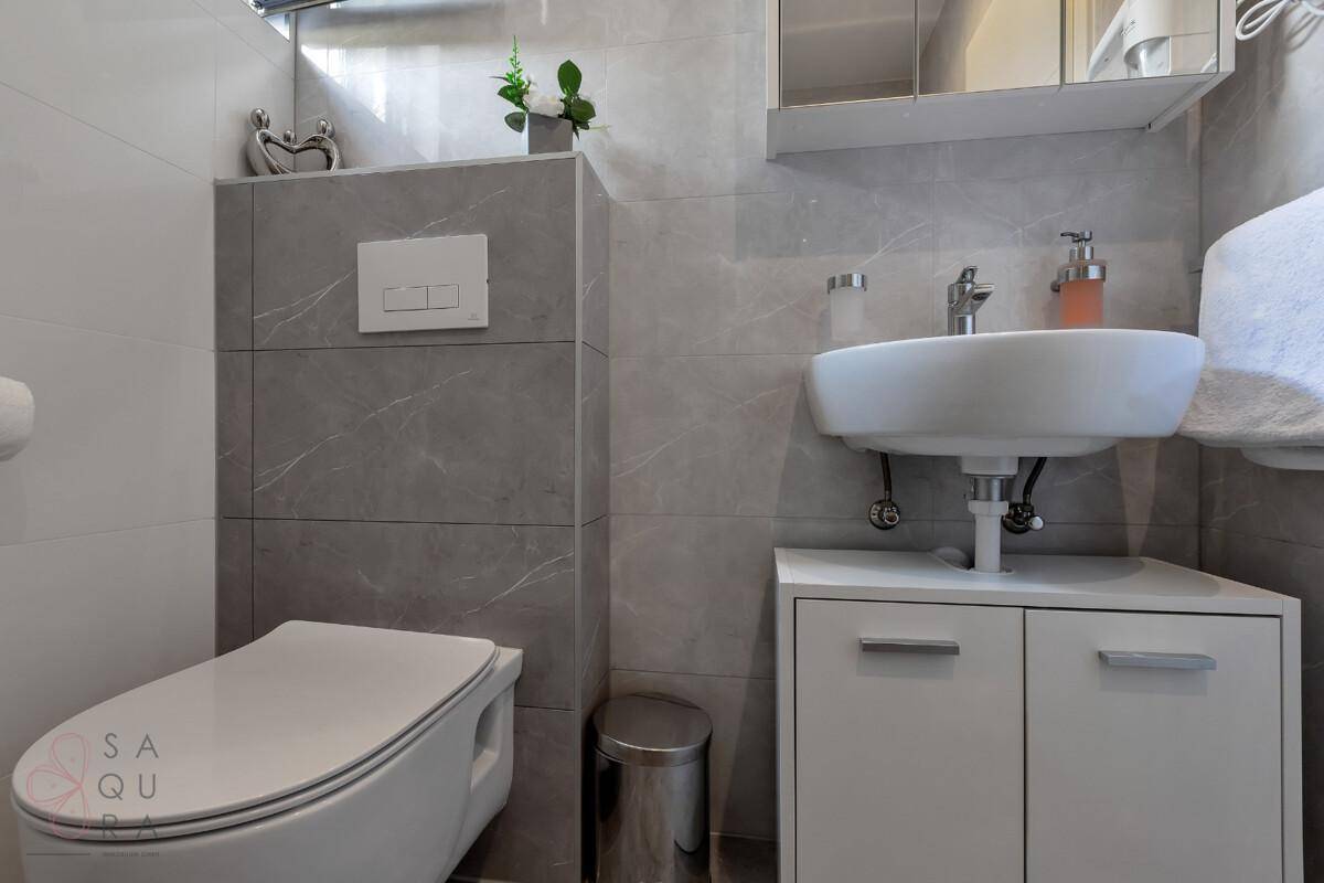 Modernes Gäste-WC mit Wand-WC, kleinem Waschbecken und grauen Fliesen.