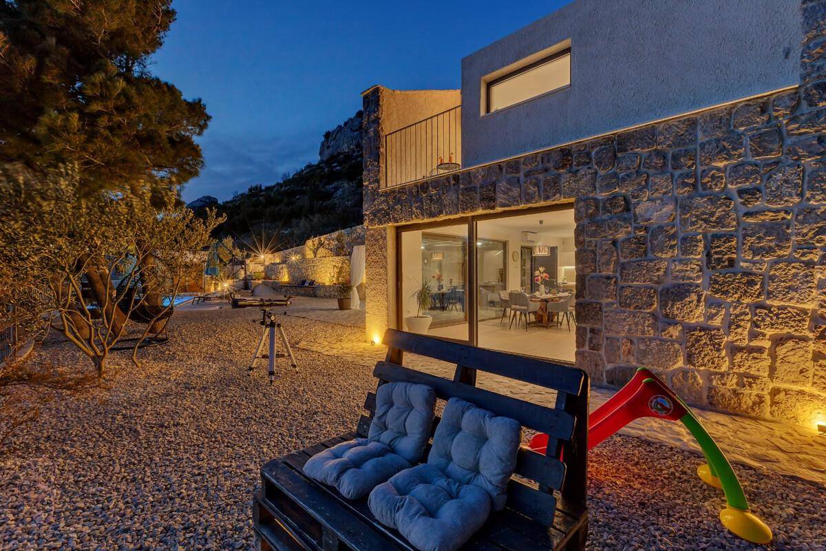 Beleuchteter Gartenbereich bei Nacht mit Sitzbank, Spielgeräten und Blick auf das Haus.