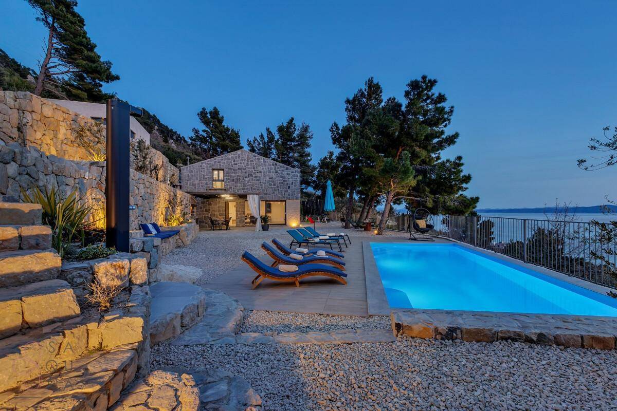 Beleuchteter Poolbereich bei Nacht mit Steintreppen und Meerblick, umgeben von mediterraner Vegetation.