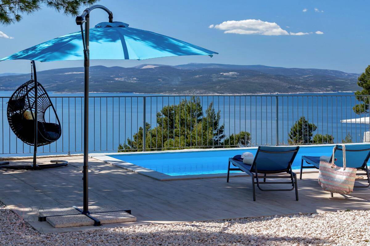Sonniger Poolbereich mit Liegestühlen und blauem Sonnenschirm, ideal zum Entspannen mit Meerblick.