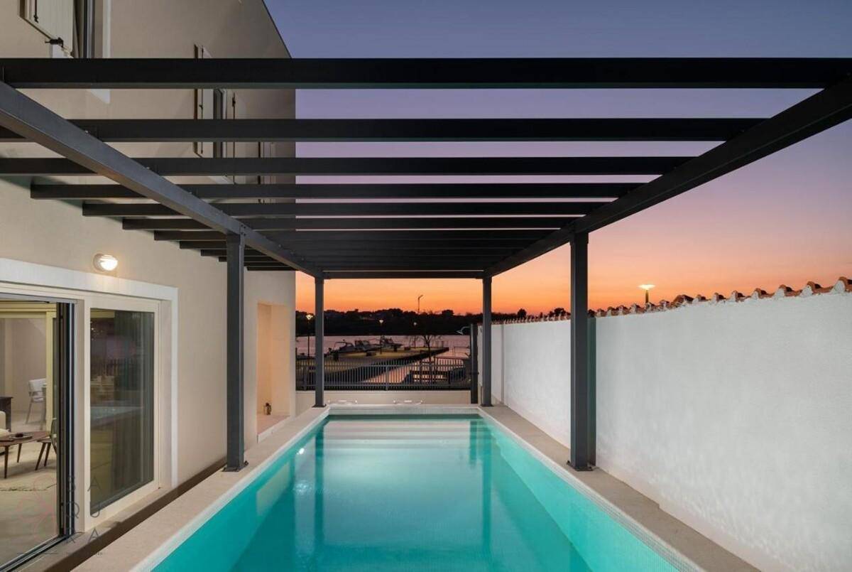 Beleuchteter Poolbereich unter einer Pergola bei Sonnenuntergang mit Blick auf das ruhige Wasser.