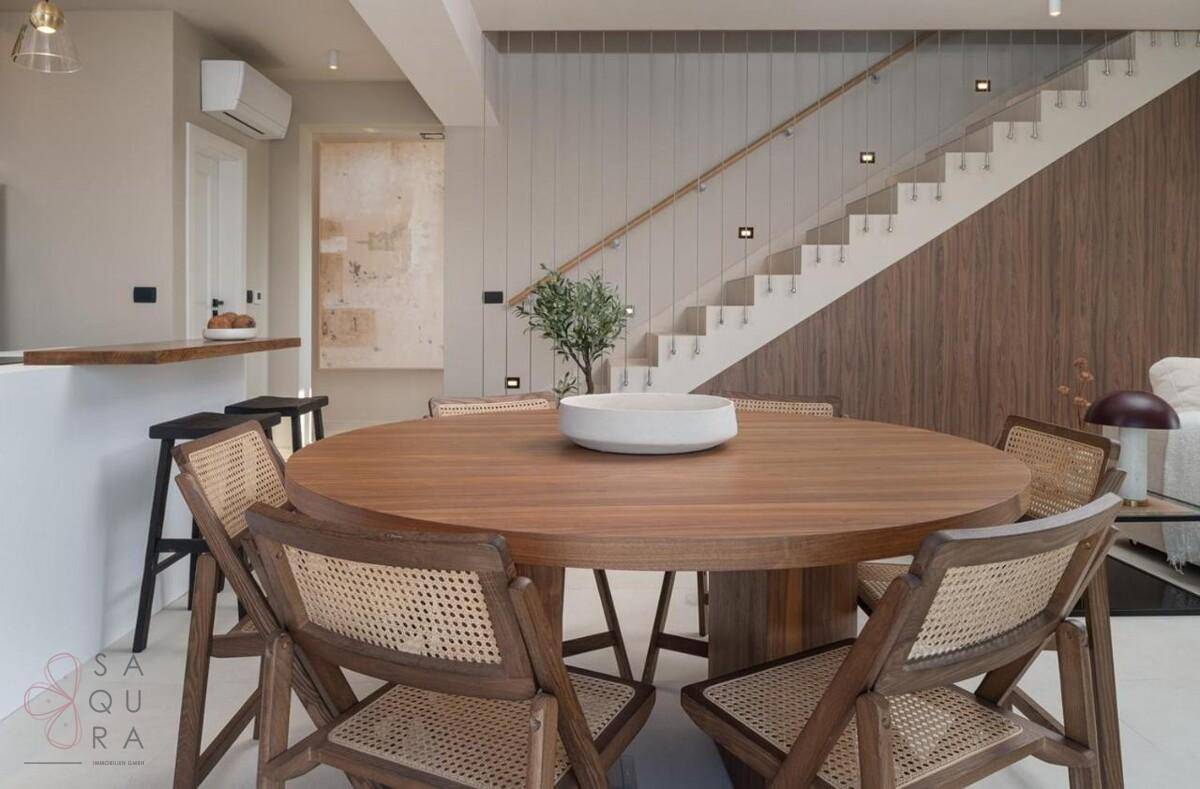 Eleganter Essbereich mit rundem Holztisch und moderner Treppe im Hintergrund.