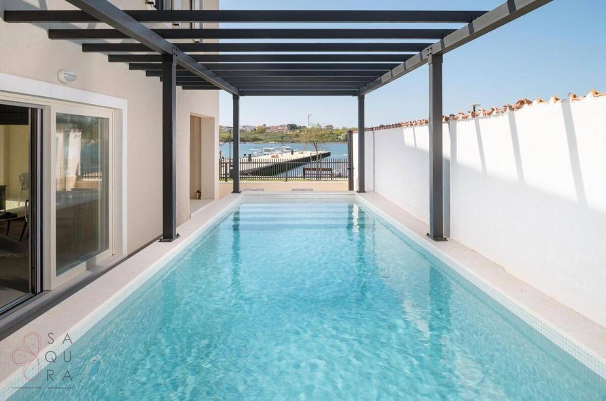 Heller Poolbereich unter einer Pergola mit direktem Blick auf das glitzernde Wasser.