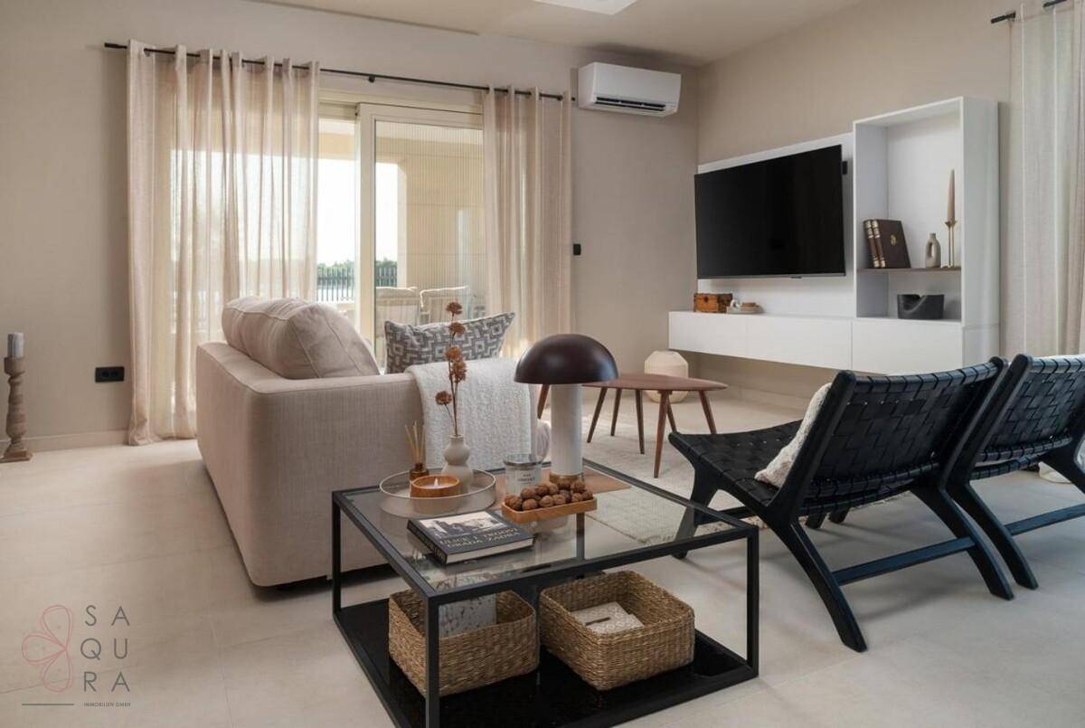 Helles Wohnzimmer mit modernem Design, beigem Sofa, Fernseher und Zugang zu einer Außenterrasse.